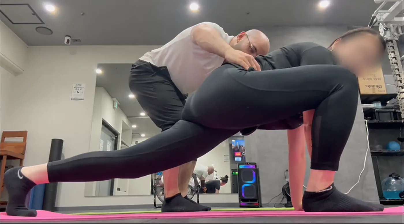 Samuel guides a client's hip position during a deep kneeling lunge stretch on a mat at a rental gym.