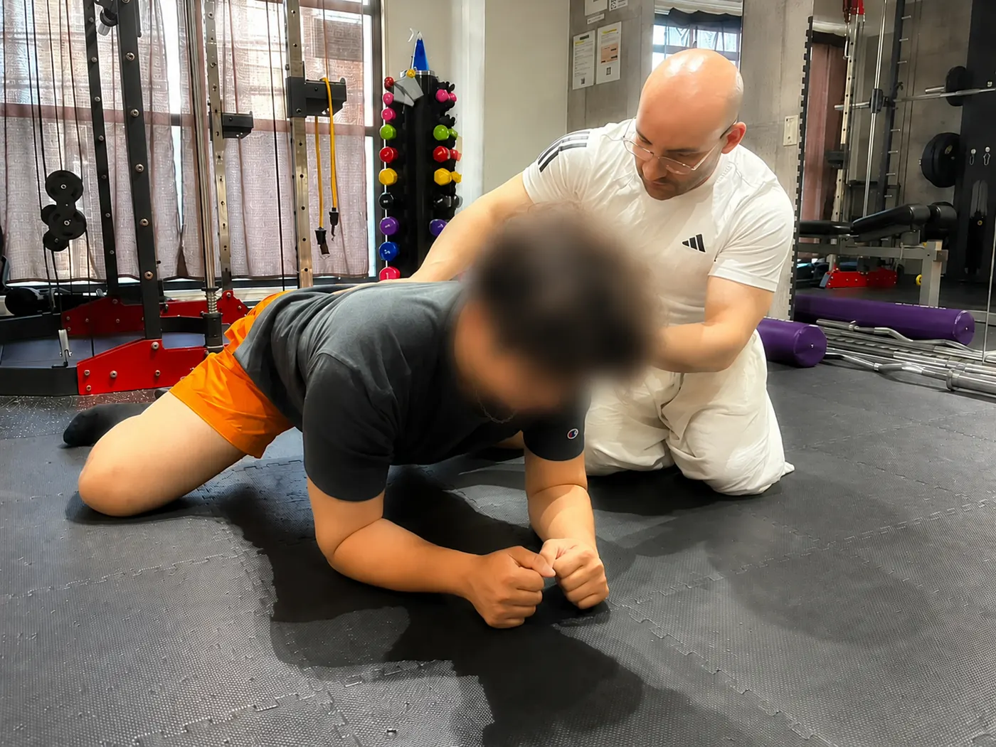 Personal trainer Samuel Ancora kneels beside a client and guides their back position during a forearm plank at a rental gym in Osaka.