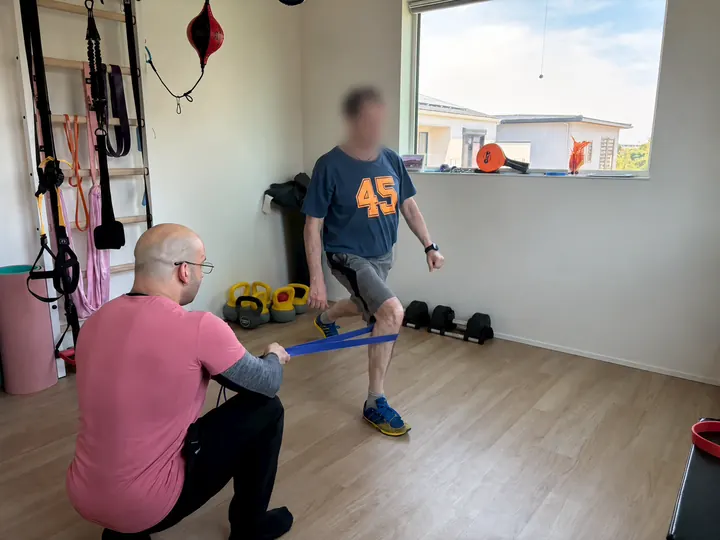 Samuel kneels and holds a resistance band around a client's knee while the client steps into a lunge.