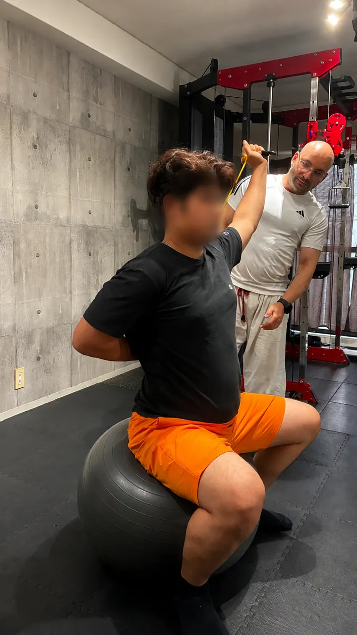 Samuel watches closely as a client seated on an exercise ball pulls a resistance band overhead.