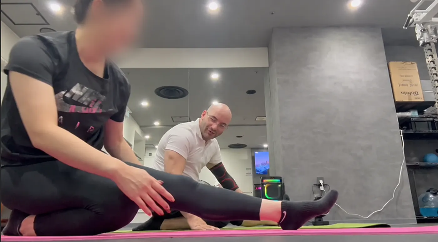 Samuel smiles and explains the next step while a client sits on a mat stretching one leg forward.